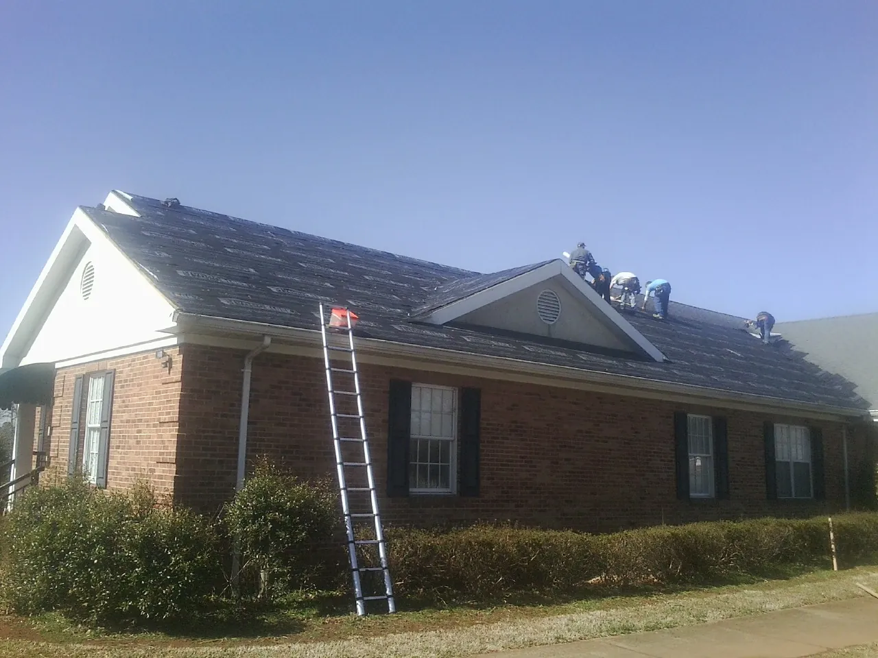 Crew tearing shingles off a brick ranch roof with a ladder set — Emergency Roof Repair work by our crew in Volga