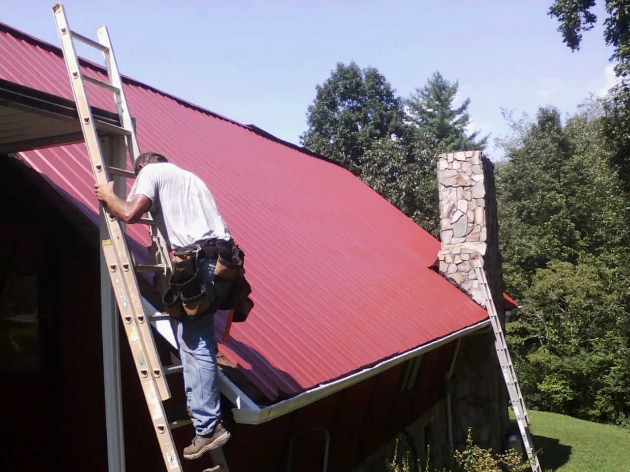 Roofer on a ladder beside a red metal roof and stone chimney — Soffit & Fascia Repair by Roofing Brothers in Volga